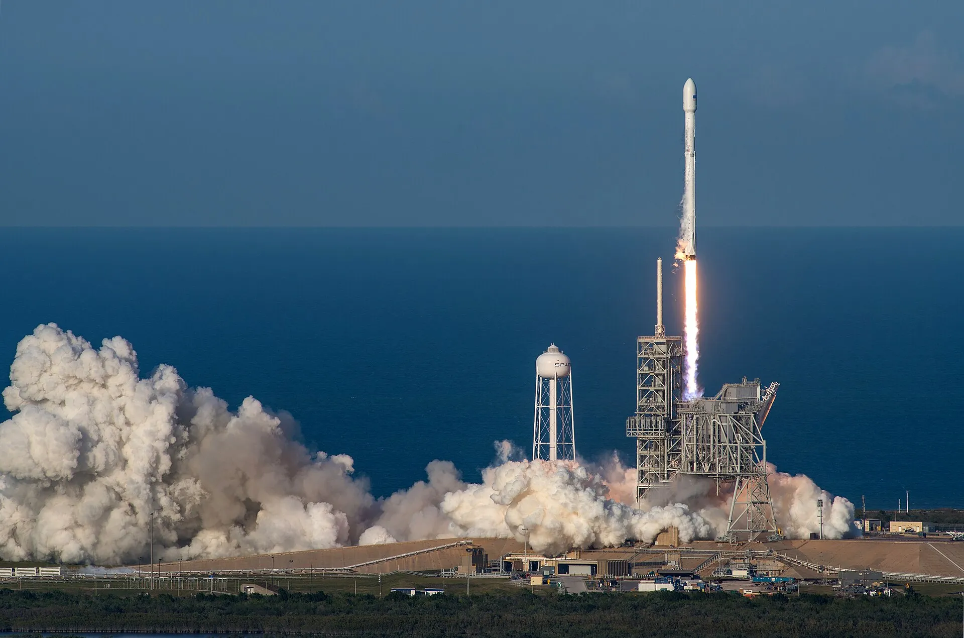 A SpaceX Falcon 9 rocket lifts off on a column of flame against a clear sky.