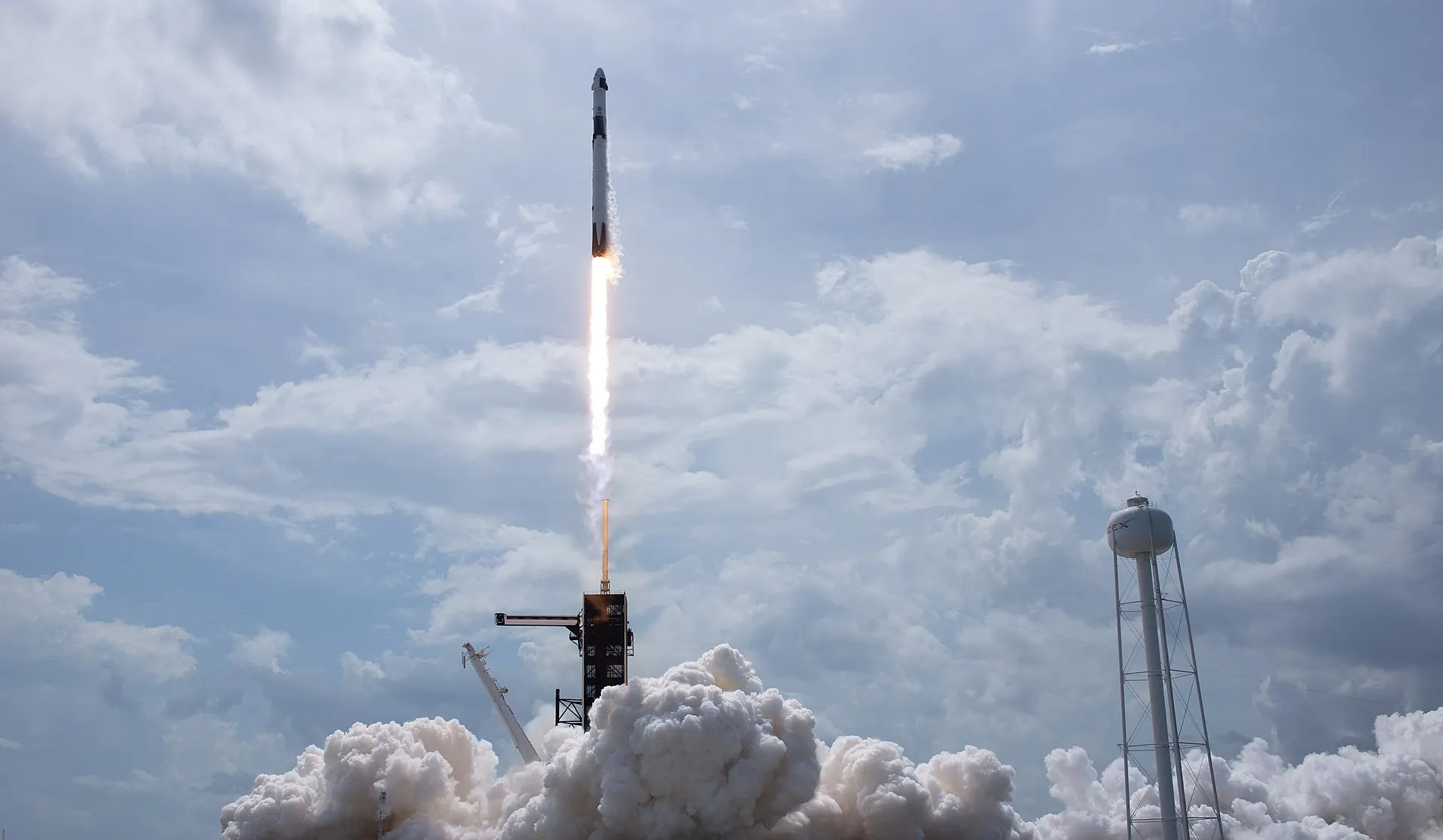 A Falcon 9 rocket lifts off from a launch pad, trailing a bright column of exhaust and smoke into a clear sky.