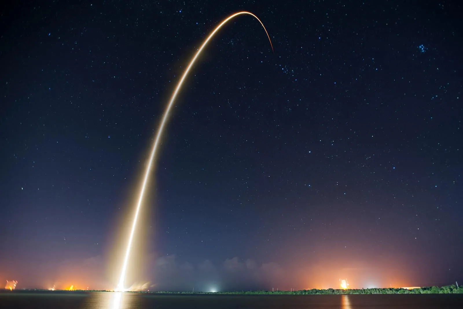A long-exposure photograph of a SpaceX rocket ascending on a bright arc of flame against a starry night sky, with the booster separation point visible as a brief second flash
