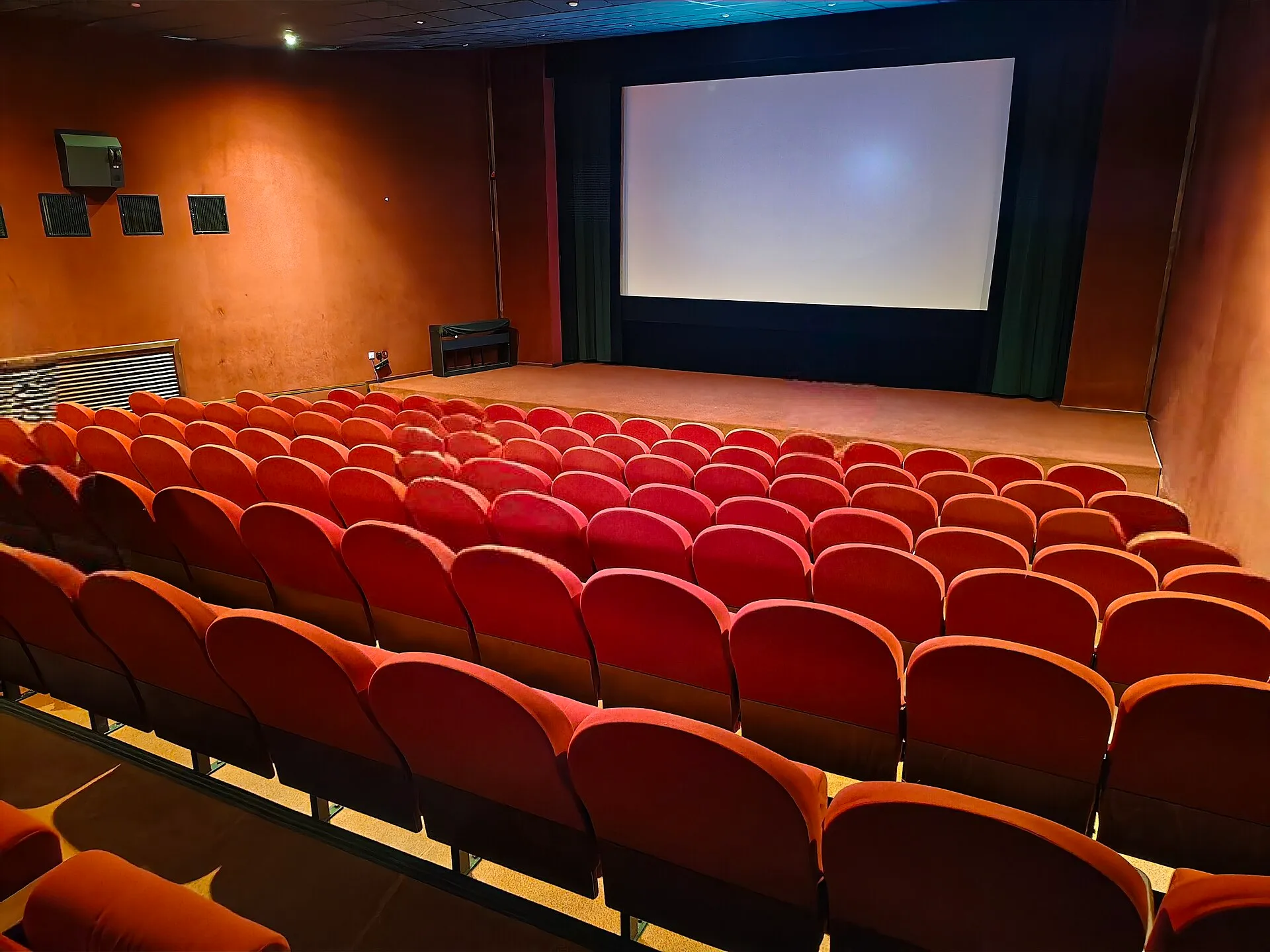 The interior of a movie theater auditorium with rows of empty red seats facing a large blank cinema screen.