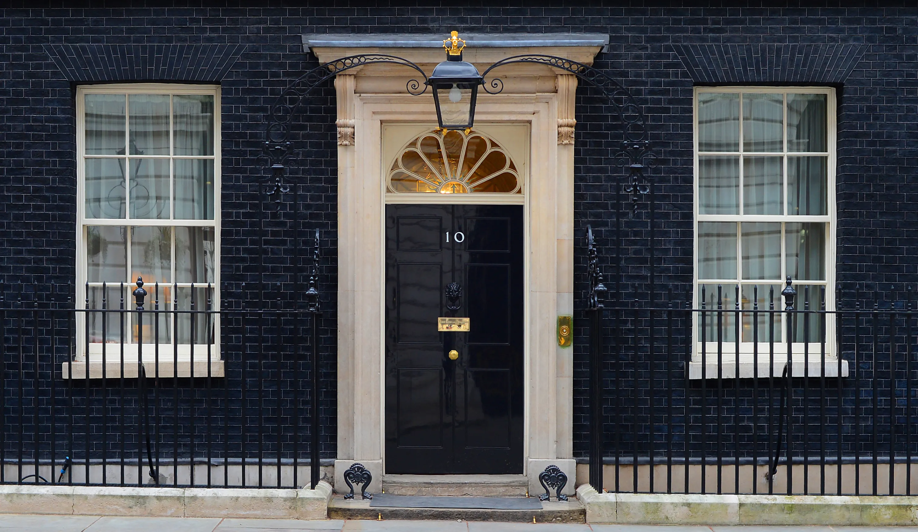 The black front door of 10 Downing Street, marked with the number 10, flanked by black iron railings and a lantern.