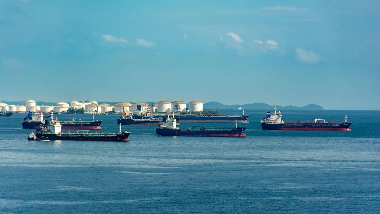 A fleet of oil tankers and storage tanks under a clear sky.