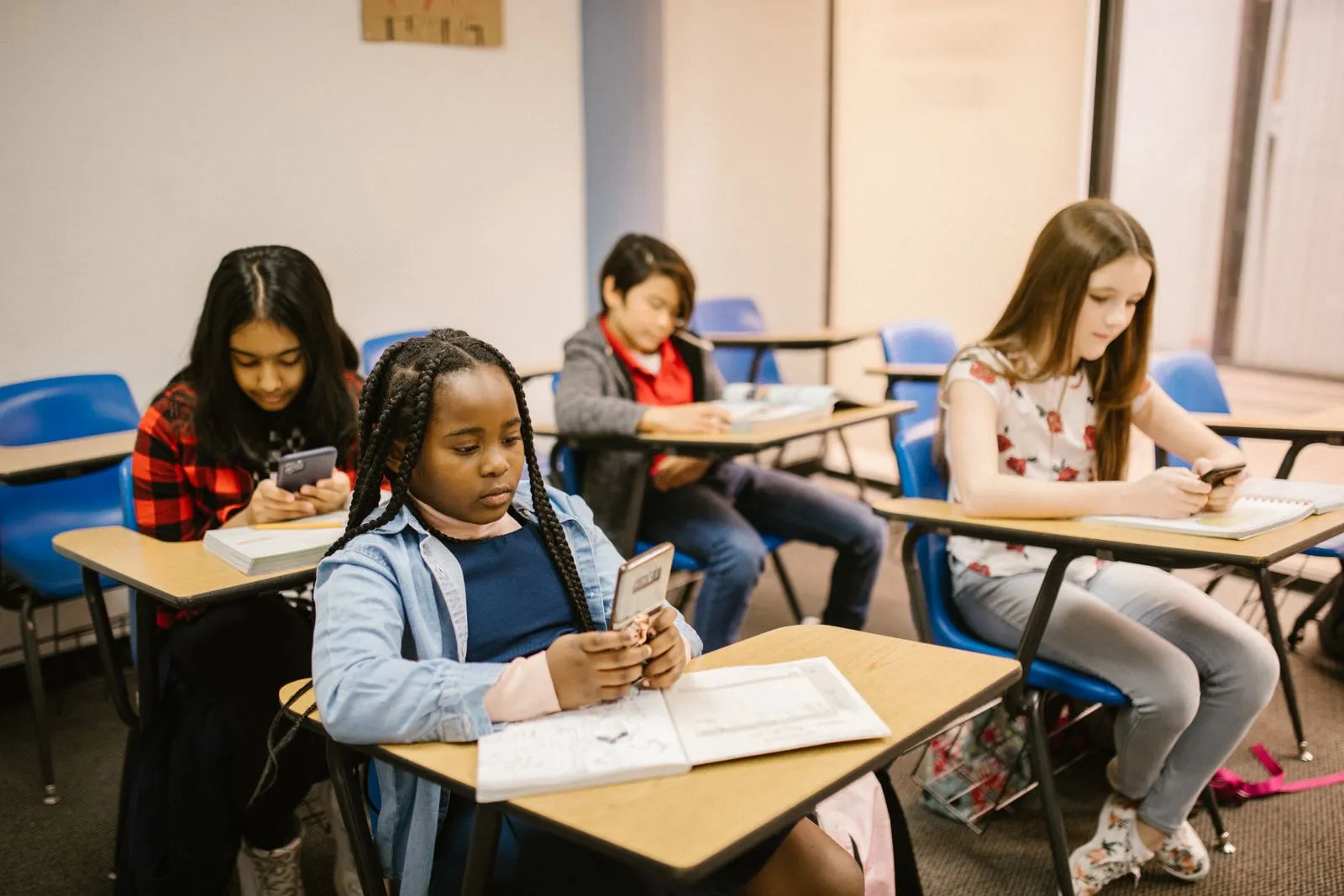 Four young students sit at classroom desks looking down at smartphones, with open notebooks and textbooks in front of them.