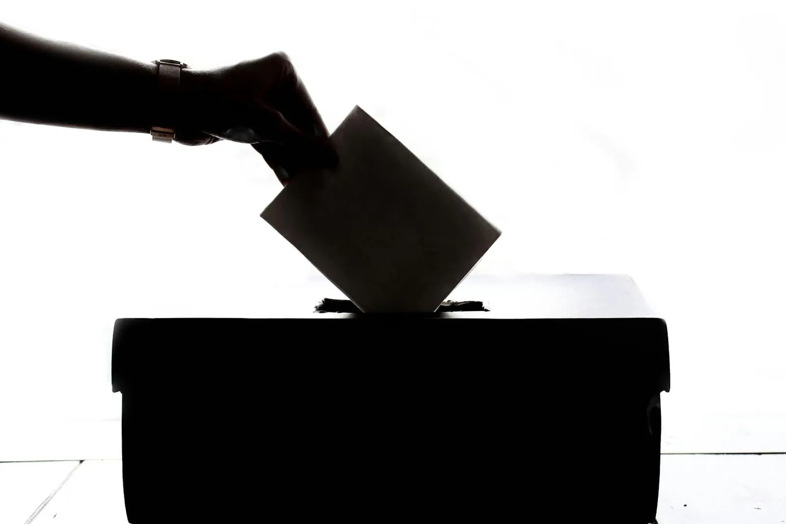 A silhouetted hand places a folded paper ballot into a white ballot box.