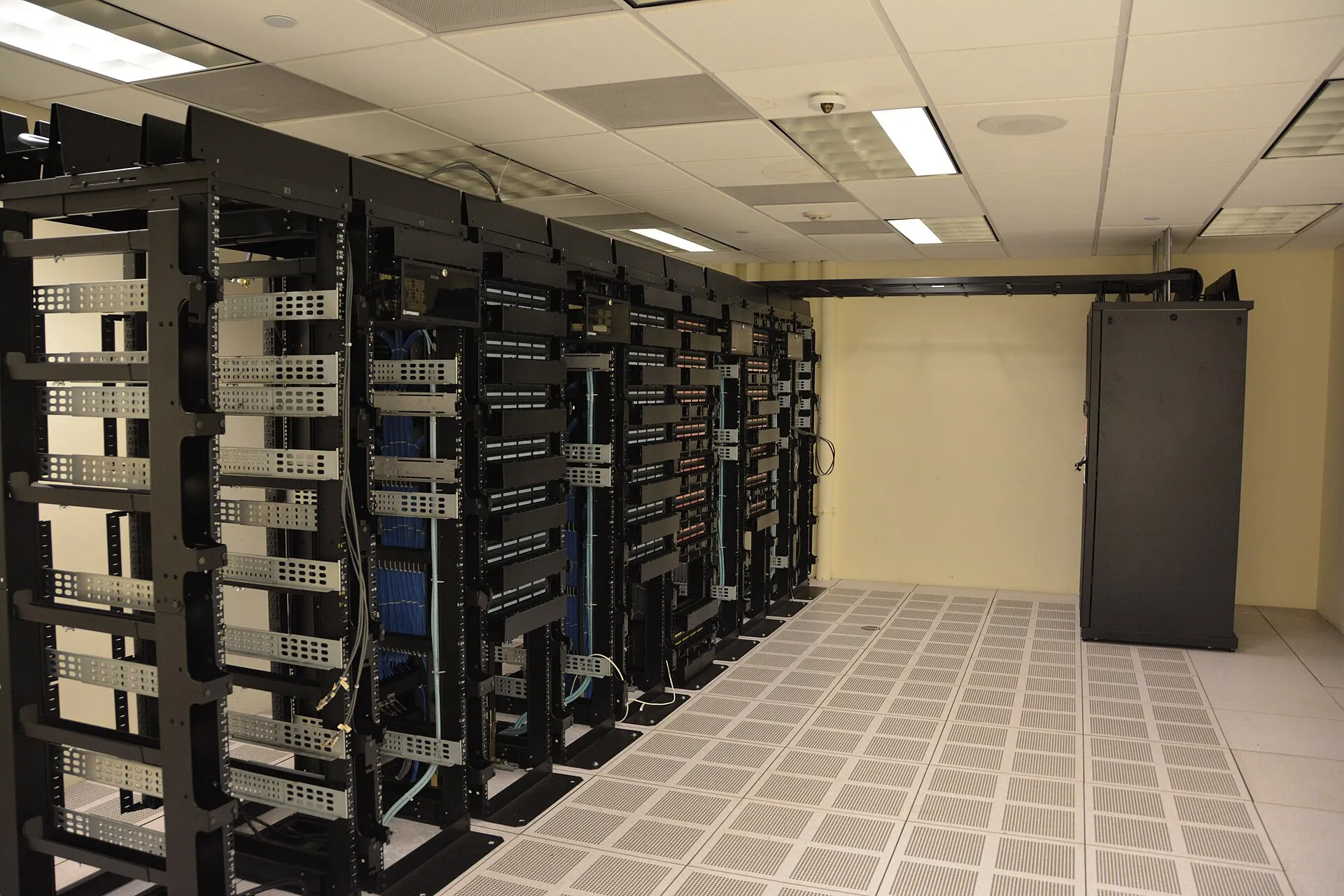 A row of dark server and networking racks with blue cabling inside a data center, on a raised perforated floor.