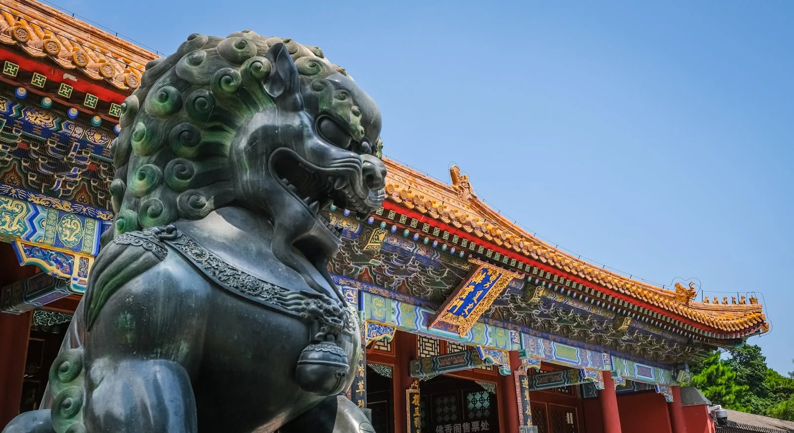 Bronze guardian lion statue outside a traditional Chinese imperial palace with colorful red and gold decorative eaves against a clear blue sky