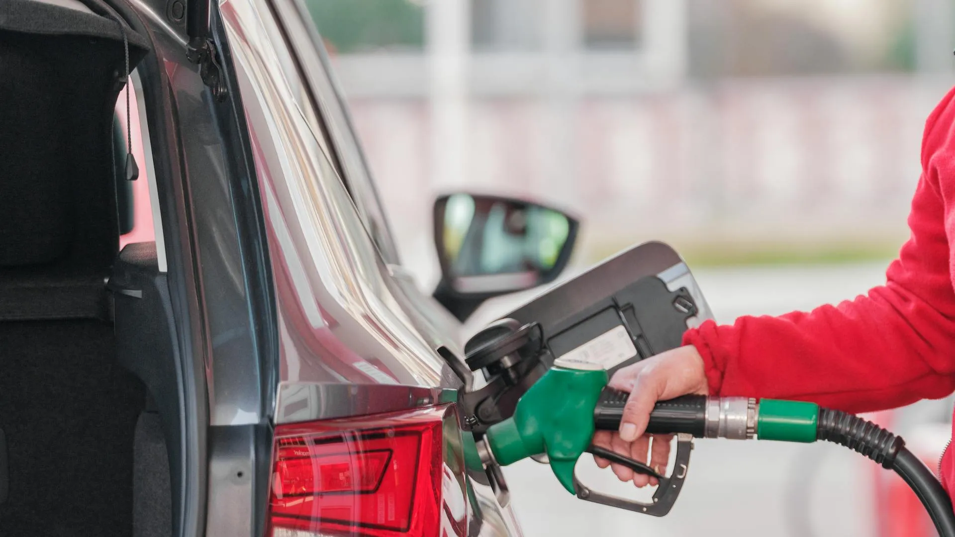 A person in a red sweatshirt gripping a green fuel nozzle while pumping gasoline into a gray car at a gas station.