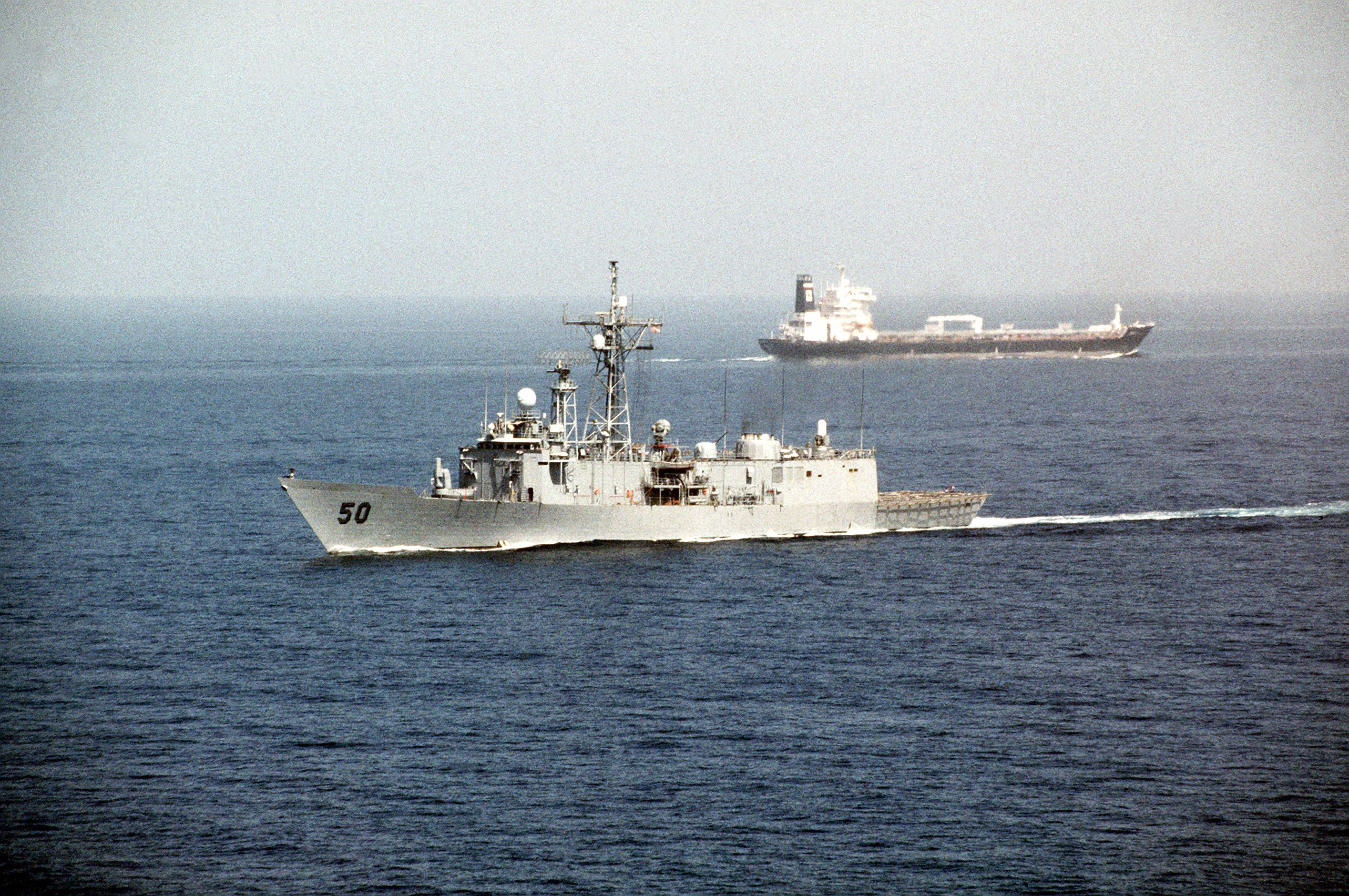 A U.S. Navy guided-missile frigate underway at sea with an oil tanker visible on the horizon behind it.