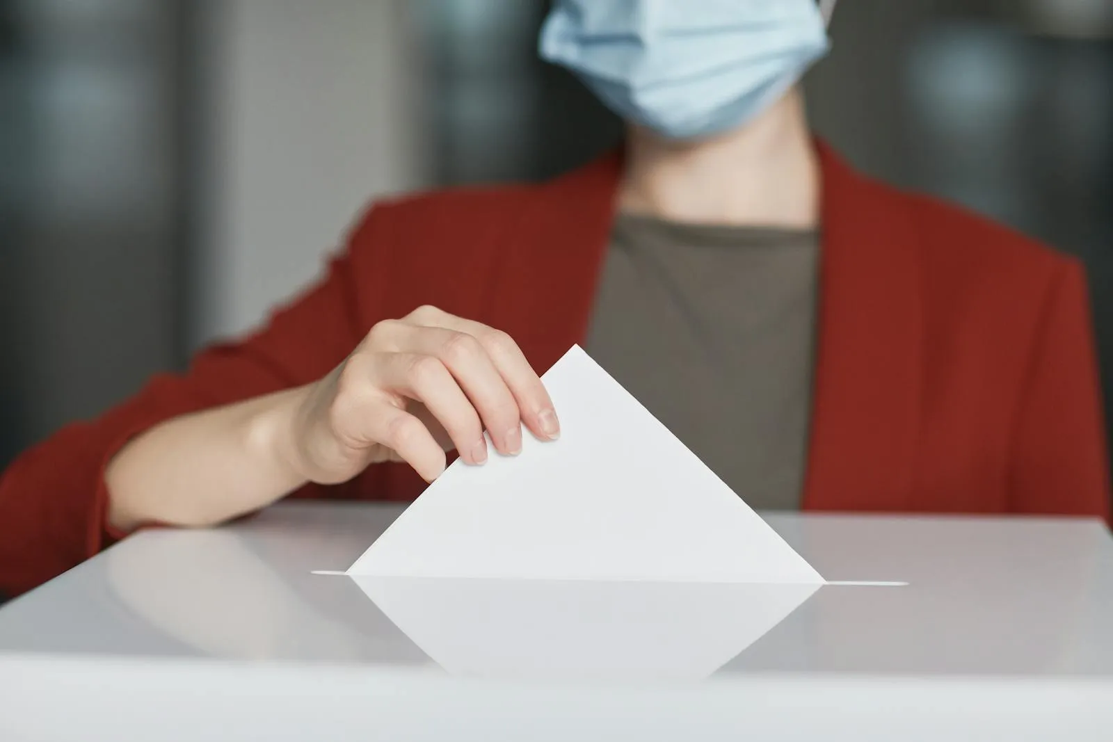 A person in a red jacket and a light blue surgical mask sliding a white folded ballot into the slot of a white ballot box.