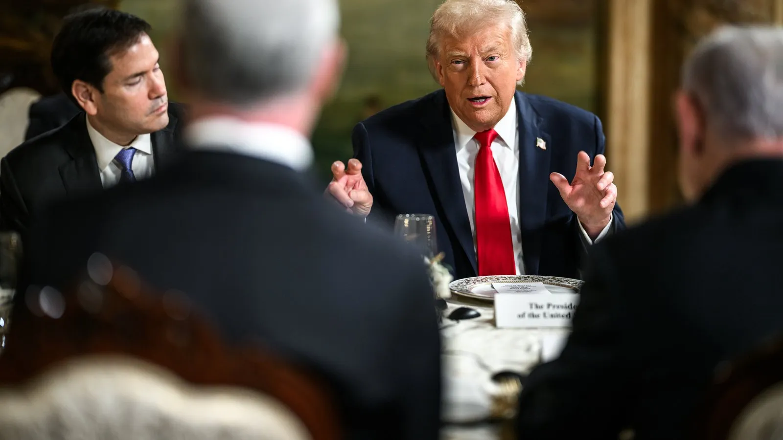 President Donald Trump speaks with hands raised at a long bilateral dining table; Secretary of State Marco Rubio sits at left, Israeli officials are visible in the foreground.