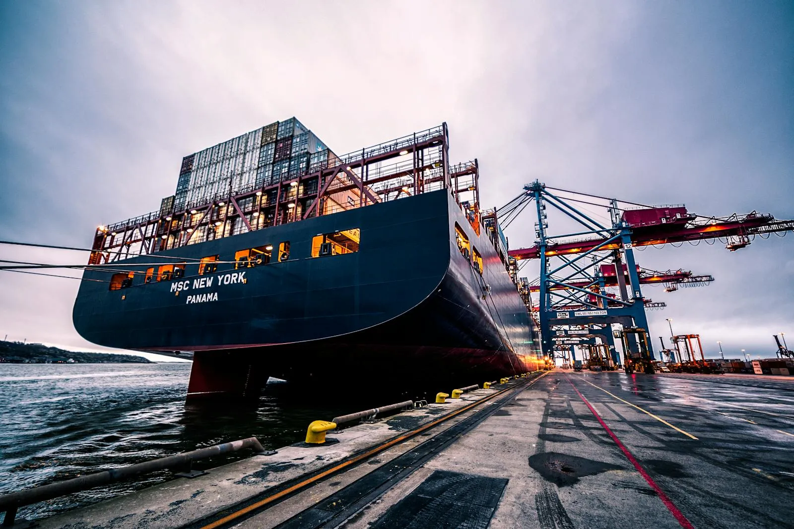 A large container ship loaded with stacked cargo containers moored at a commercial port, viewed from dock level at the bow under a cloudy sky.