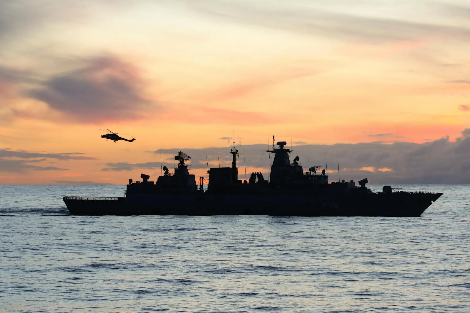 A naval warship silhouetted against an orange and gray sky on the open ocean at dusk