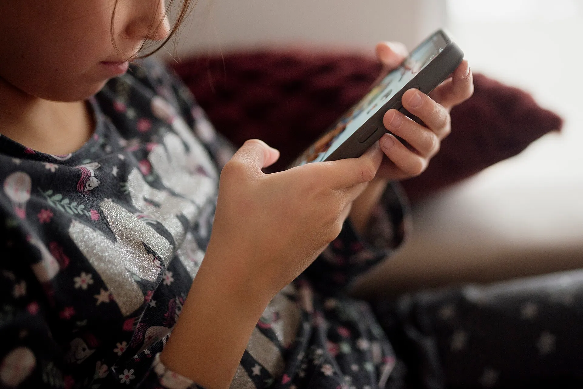 A young child sits on a couch looking down at a smartphone held in both hands.