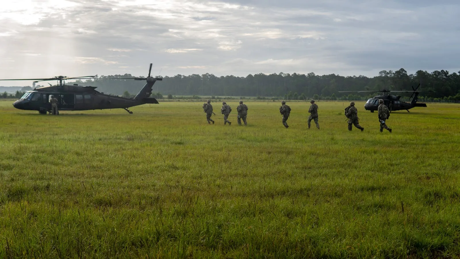 Soldiers in camouflage gear walking across a green field between two military helicopters under an overcast sky