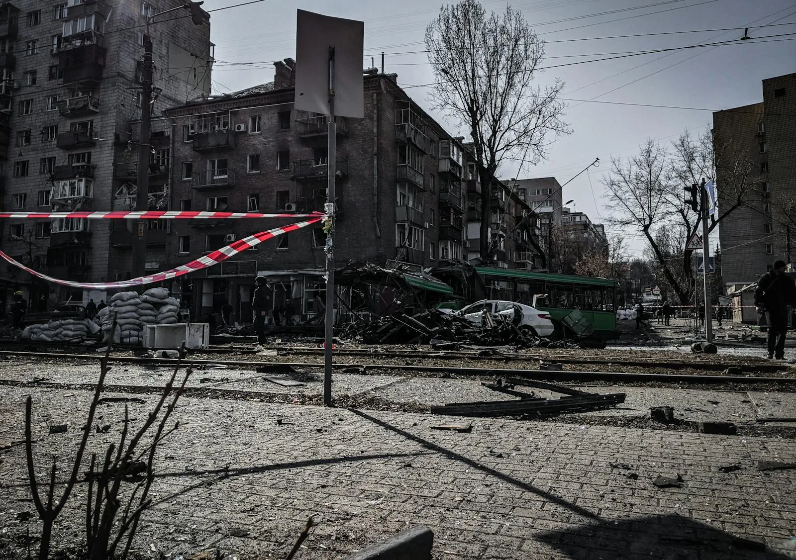 A destroyed trolley bus and damaged apartment buildings in a Ukrainian city street, cordoned off with red and white police tape after a strike