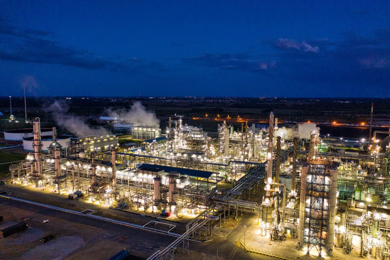 An oil refinery lit up at night, with storage tanks, towers, and pipework glowing against a dark sky.