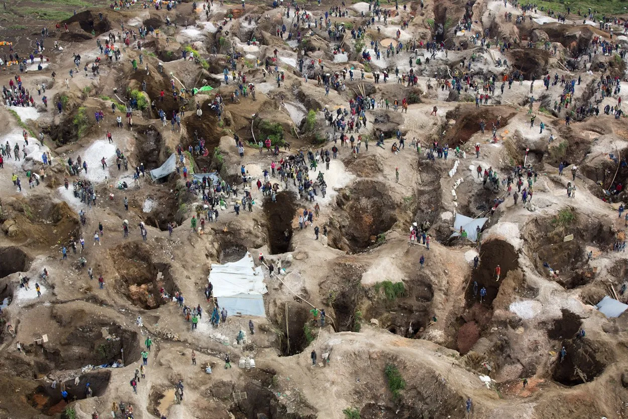 Aerial view of an open-pit artisanal mine where hundreds of workers dig across muddy terraced pits and shafts.