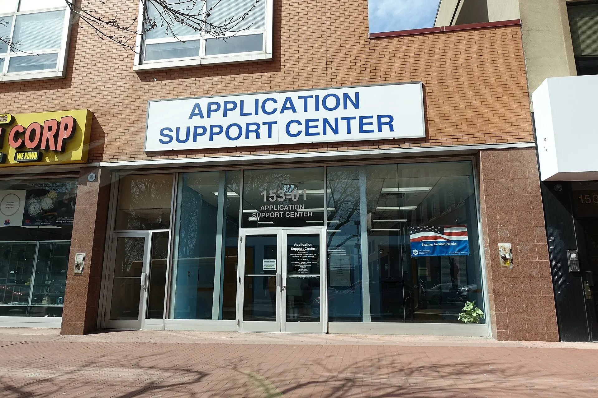 Exterior facade of a USCIS Application Support Center building at a street corner in Queens, New York.