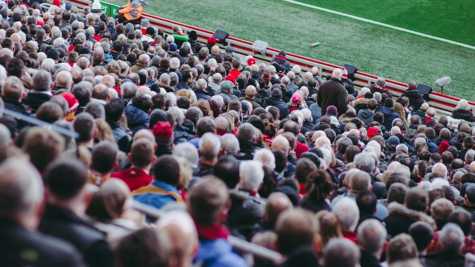 Densely packed crowd of spectators filling the stands at a soccer stadium, with a strip of green pitch visible above.