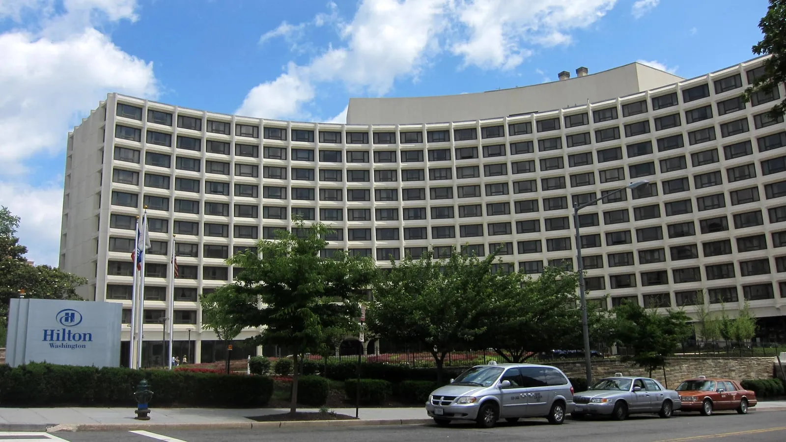 Daytime exterior view of the Washington Hilton hotel from T Street, a tall curved modernist building with rows of balconies and a porte-cochère.
