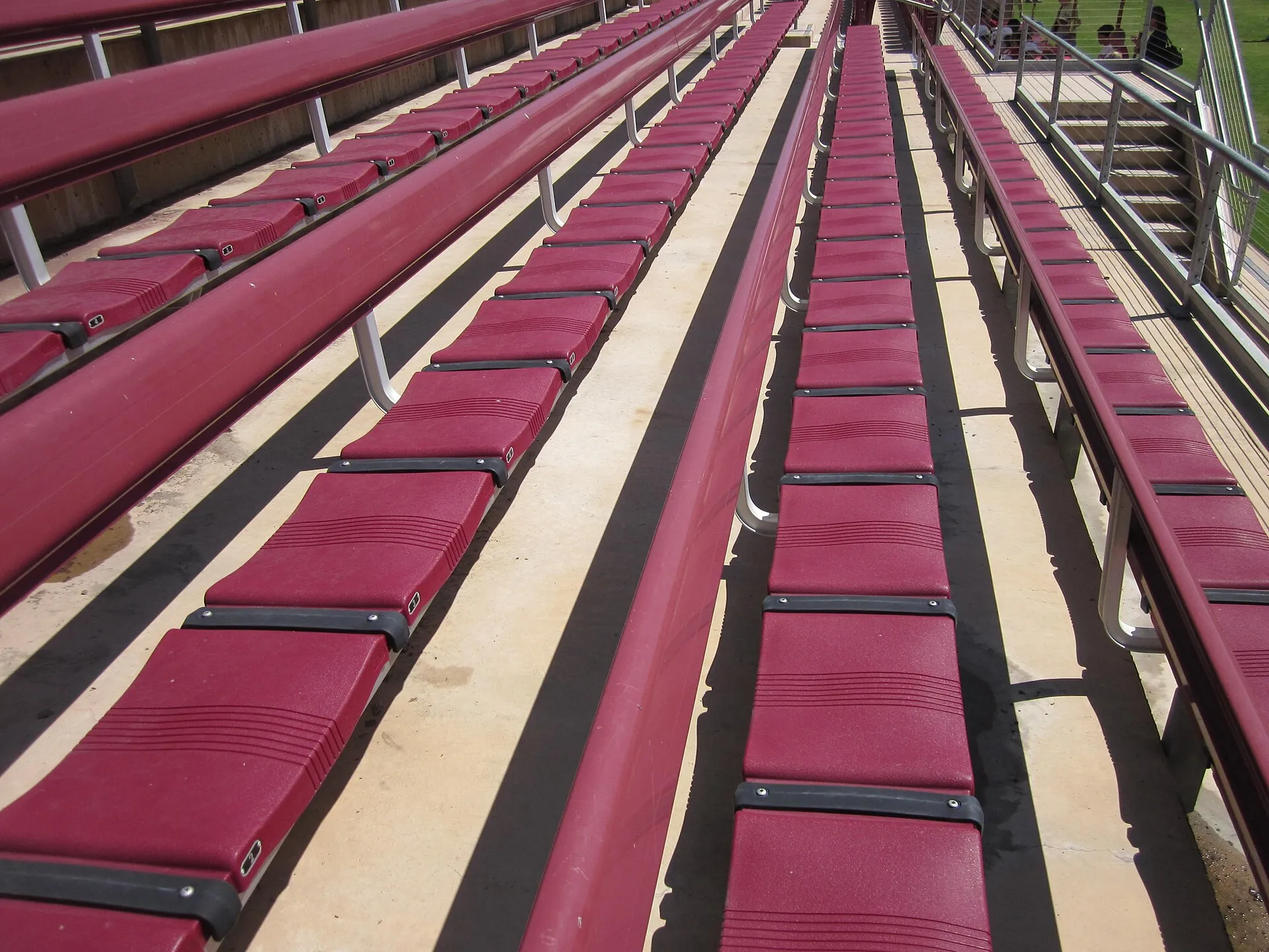 Empty rows of dark-red bench seats with metal railings in an outdoor stadium grandstand.