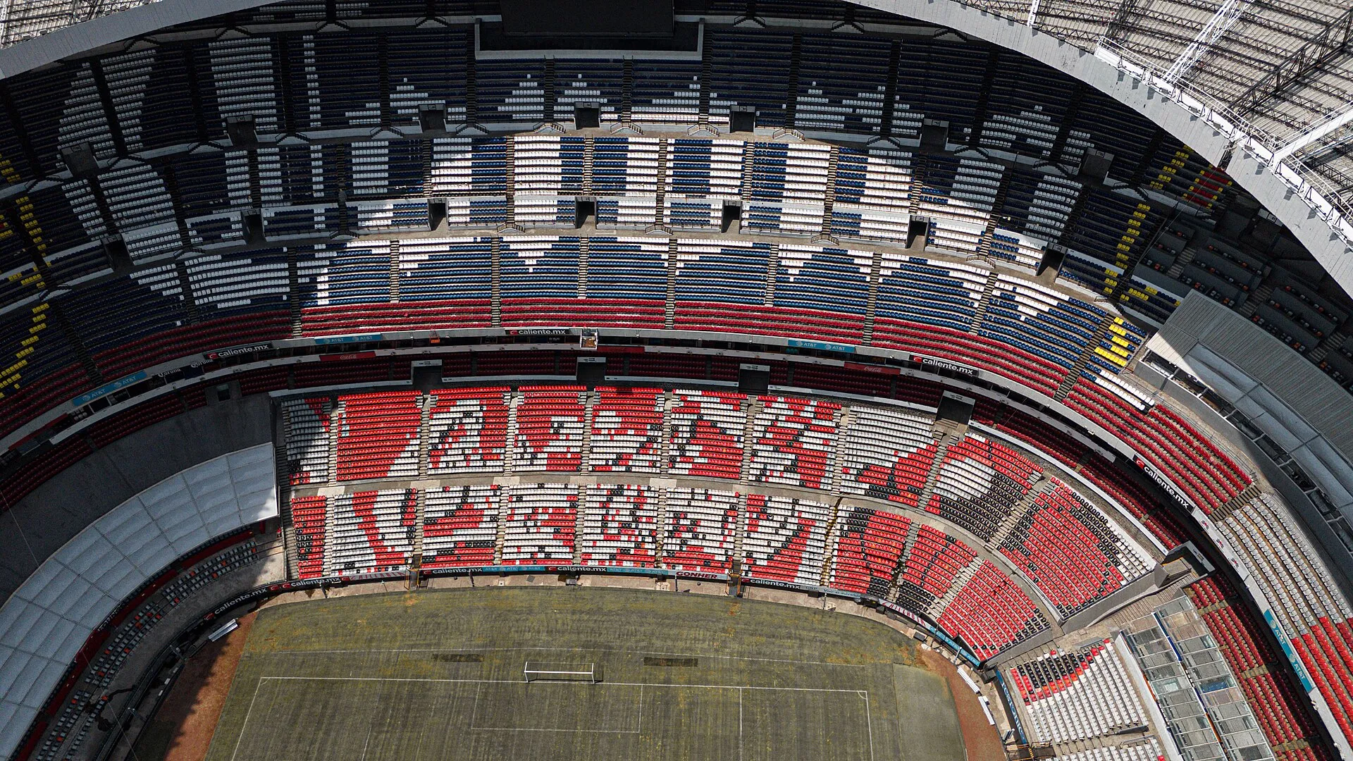 An aerial view of Estadio Azteca, a large bowl-shaped soccer stadium amid dense low-rise neighborhoods in Mexico City.