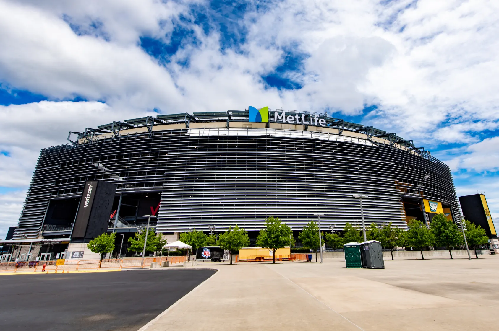 The dark louvered exterior of MetLife Stadium rising above an empty paved forecourt under a partly cloudy sky.