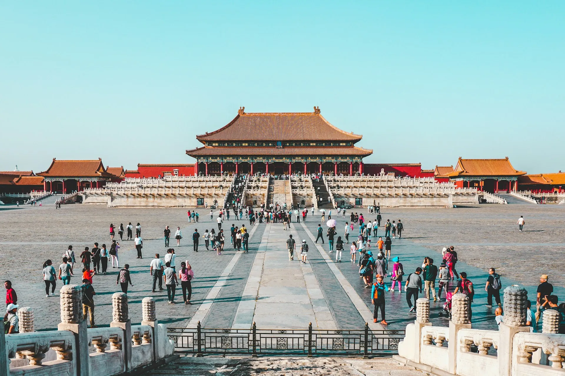 The Hall of Supreme Harmony at the Forbidden City in Beijing, with golden-tiled curved roof, red columns, and ornate eaves seen across a wide stone plaza under a clear sky, with distant figures crossing the courtyard