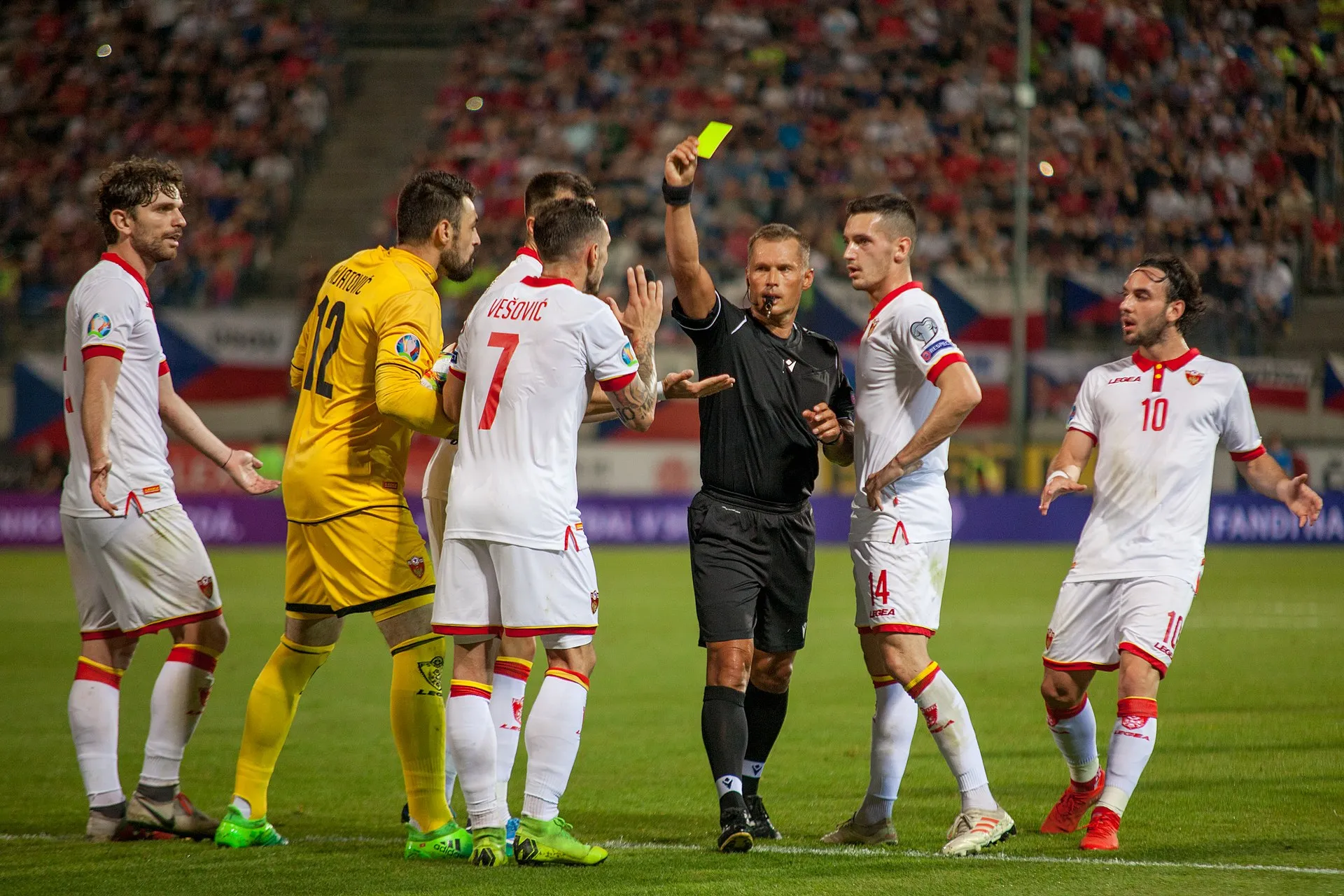 A referee in black holds up a yellow card to a group of soccer players during a floodlit match.