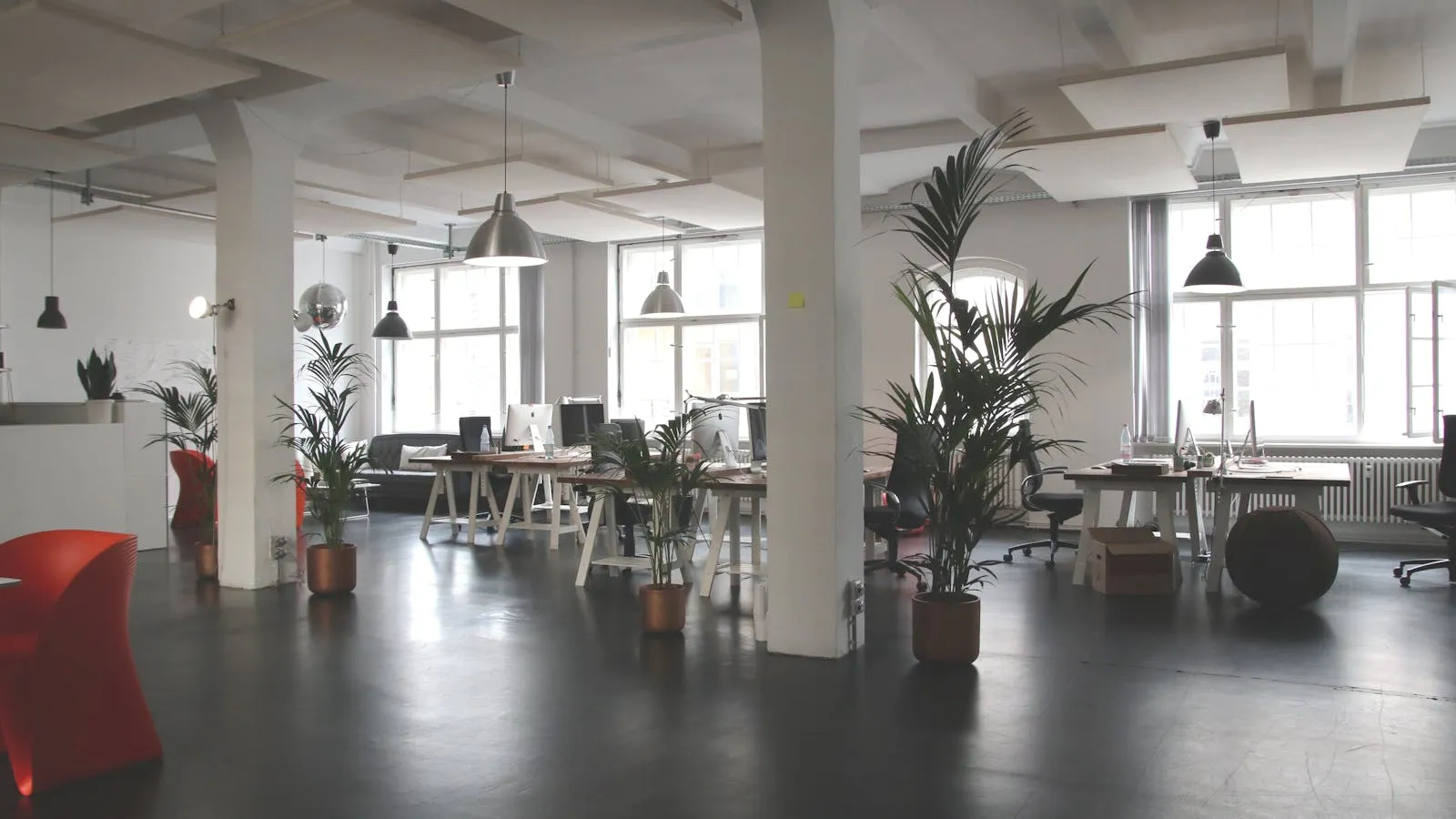 An empty open-plan office with rows of unoccupied desks, monitors, and chairs lit by daylight from large windows.